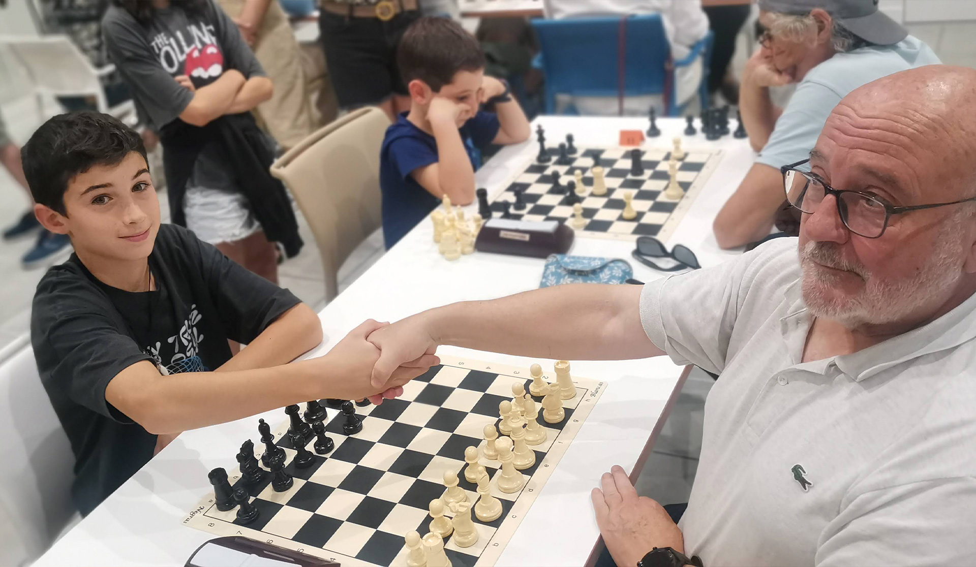 Una persona mayor estrechando la mano a un niño, sentados en una mesa con un juego de ajedrez