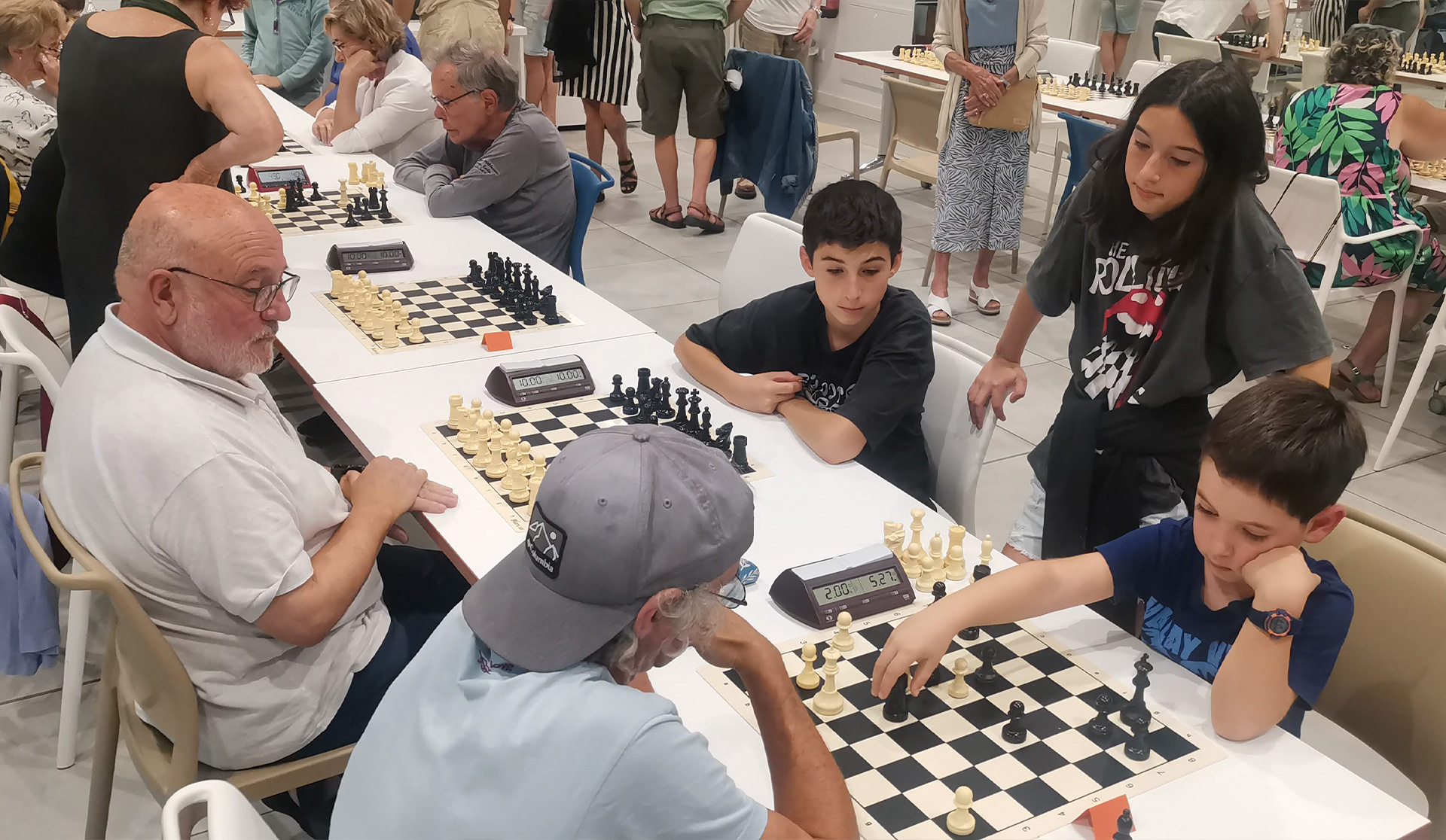 Varias mesas con gente jugando al ajedrez, mayores y niños