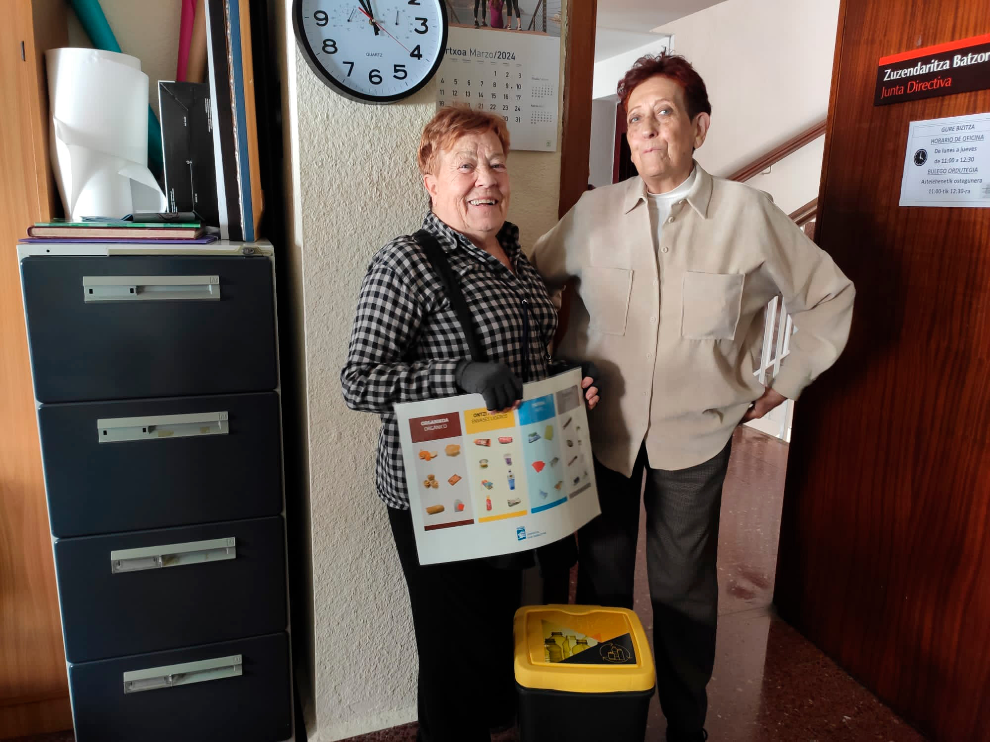 Persona sujetando un cartel con cubos de reciclaje a un lado y otra persona acompañçandola
